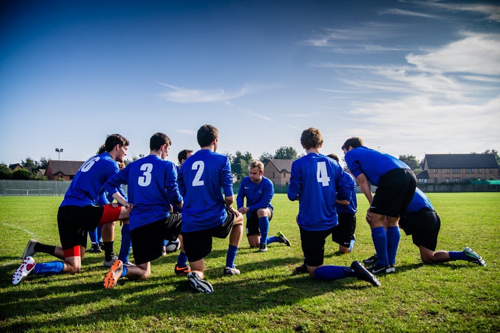 A group of male football players in blue uniforms huddle on a grassy field before a match.