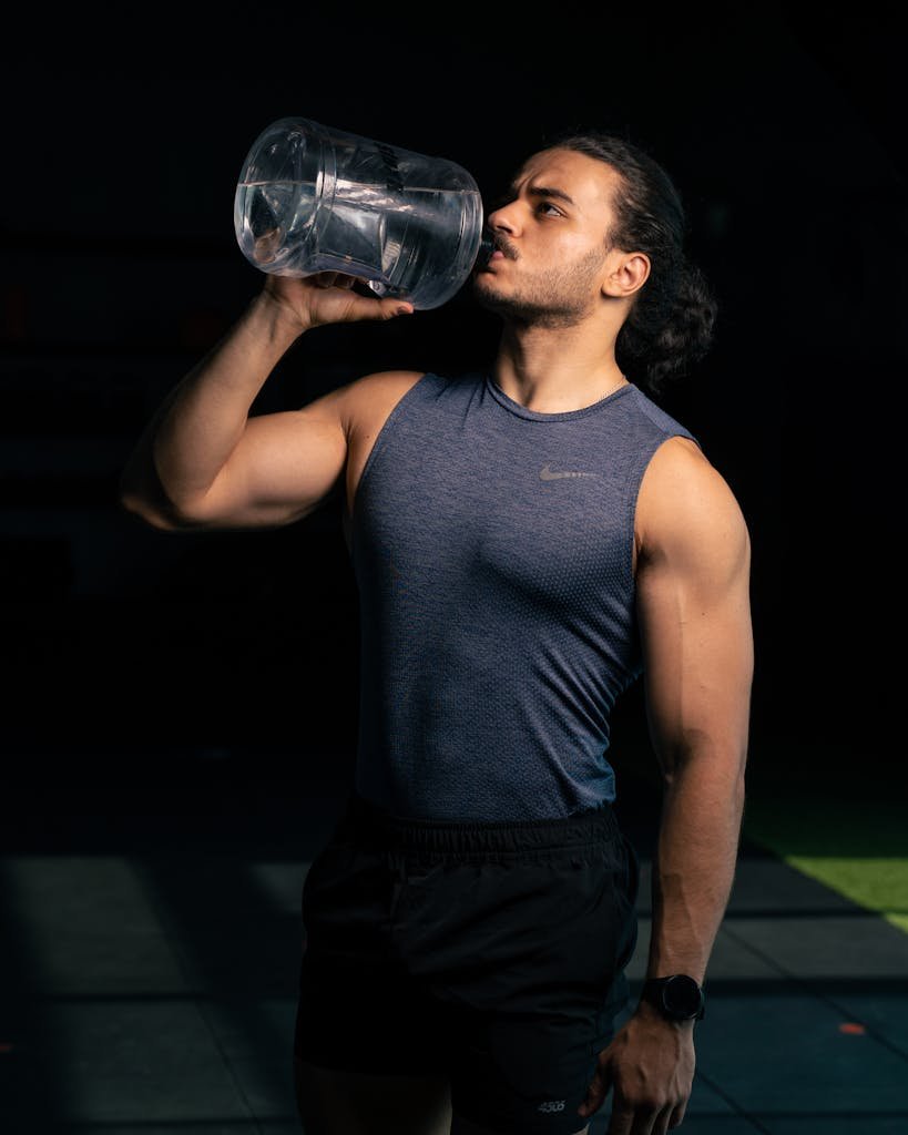 A muscular man with long hair drinks water from a large bottle post-workout.