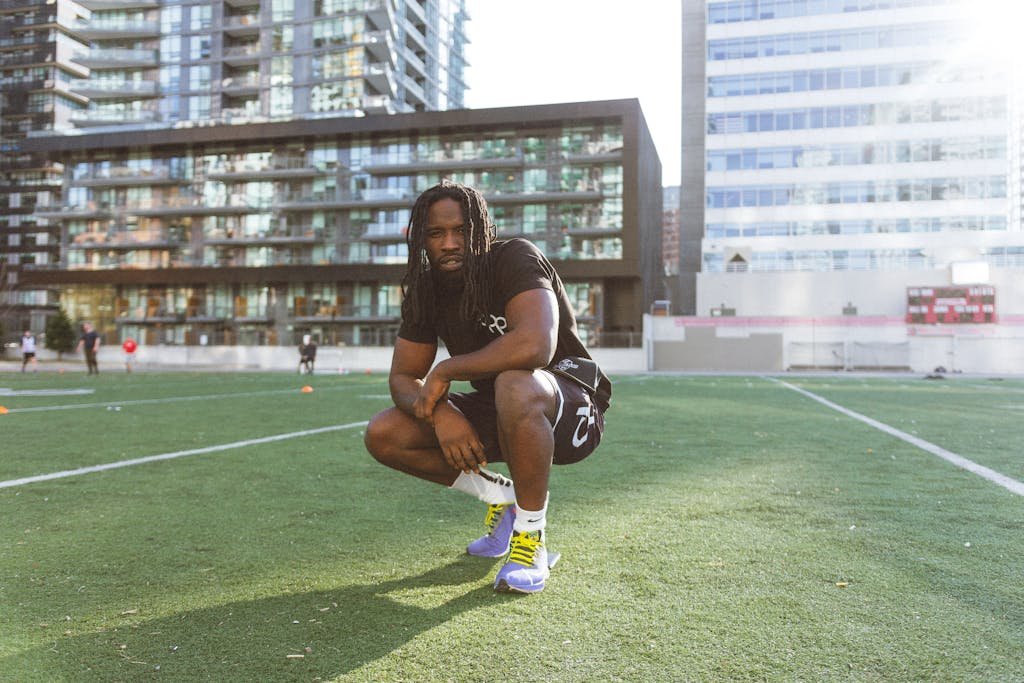 Crouching man in sportswear on a city football field with modern buildings.