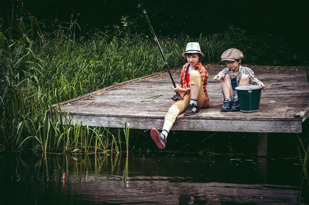 Two boys fishing on a wooden dock by a lake. They wear vintage attire and enjoy a summer day.