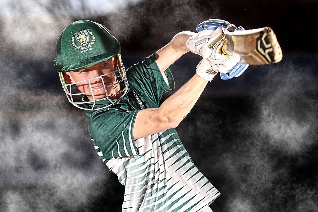 Cricket player in mid-swing with bat, wearing green uniform and helmet.