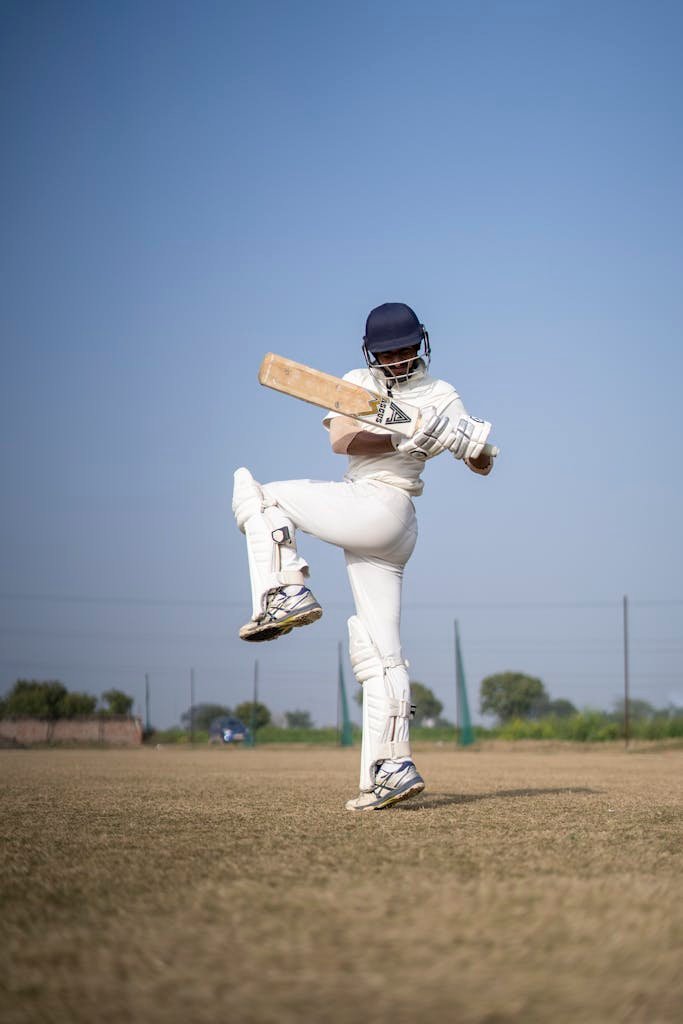 Cricket player swinging bat on a sunny field, showcasing athletic skill.