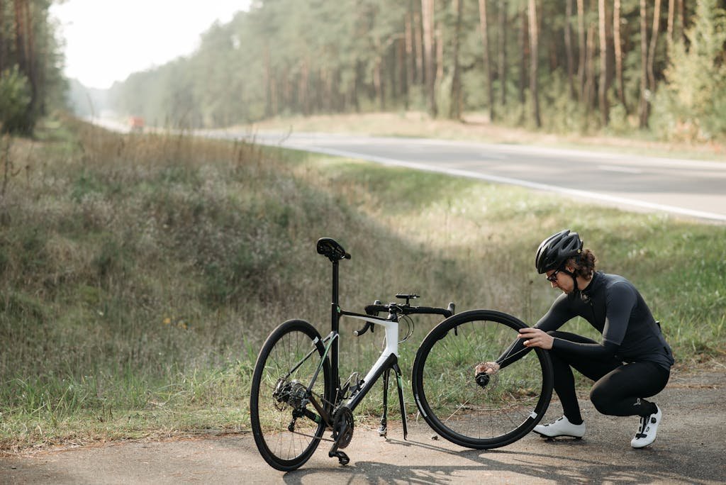 Cyclist fixing a bicycle wheel on a rural roadside surrounded by forest.