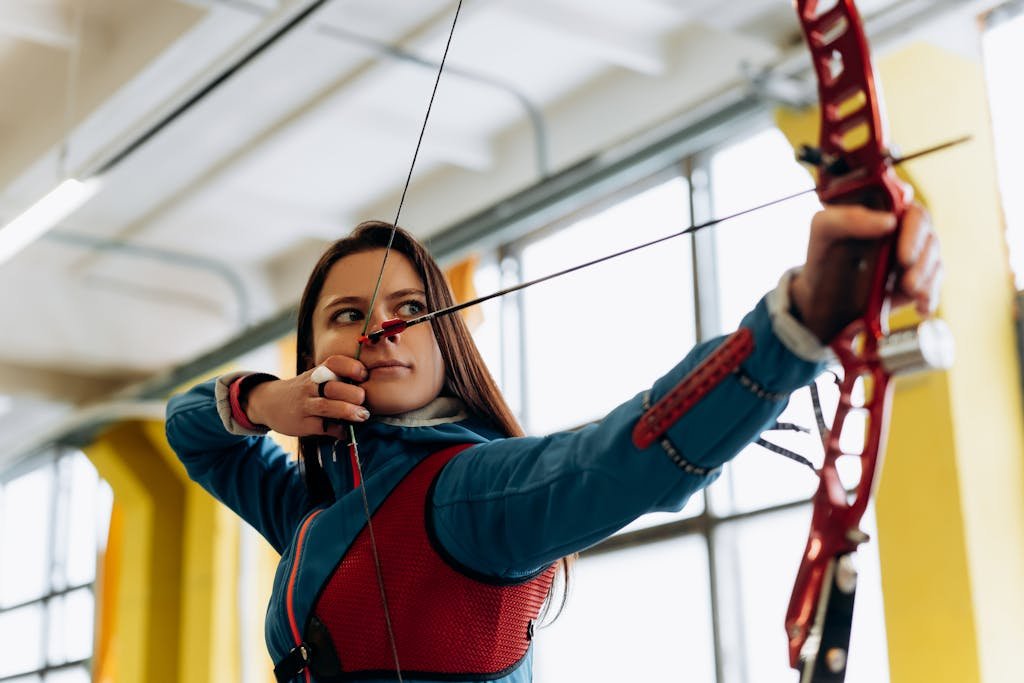 Young female archer in blue aiming a bow and arrow indoors, showcasing concentration and athleticism.