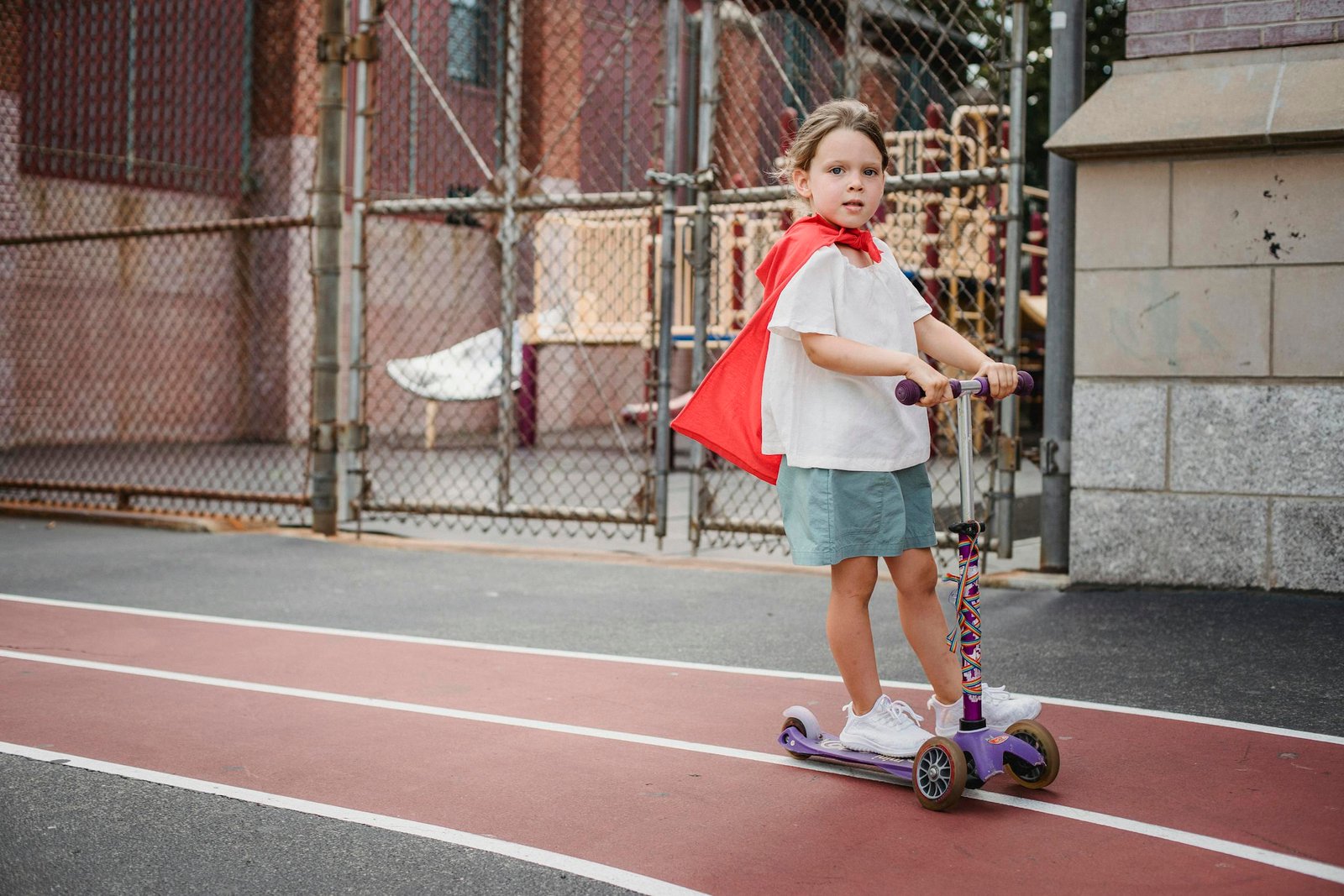 A young girl dressed as a superhero riding a kick scooter in an outdoor playground.