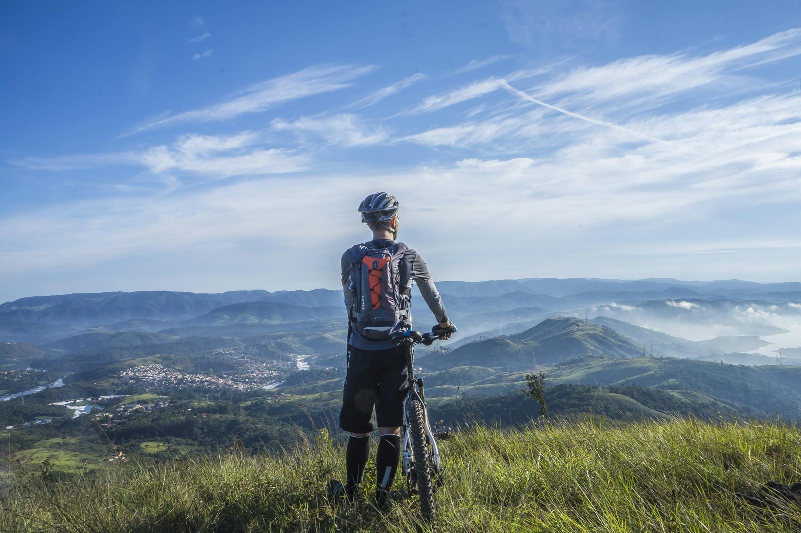 A cyclist stands on a hill, gazing at a stunning mountain landscape on a bright sunny day.