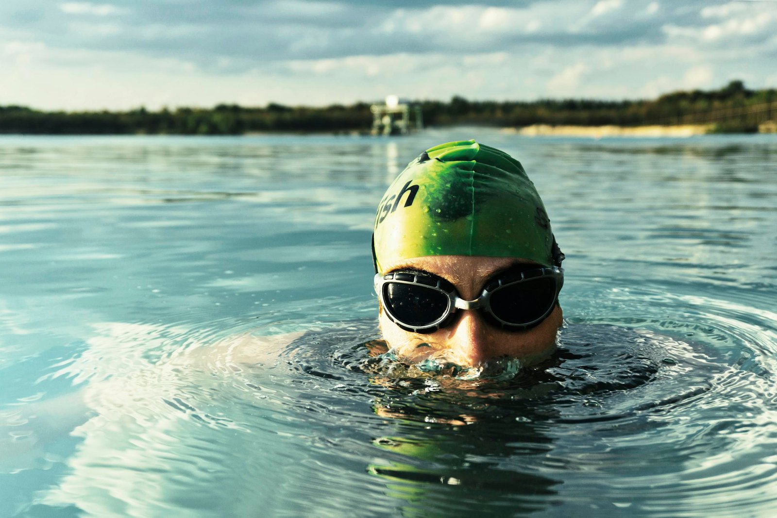 A determined swimmer in goggles and cap emerges from a tranquil lake on a sunny day.