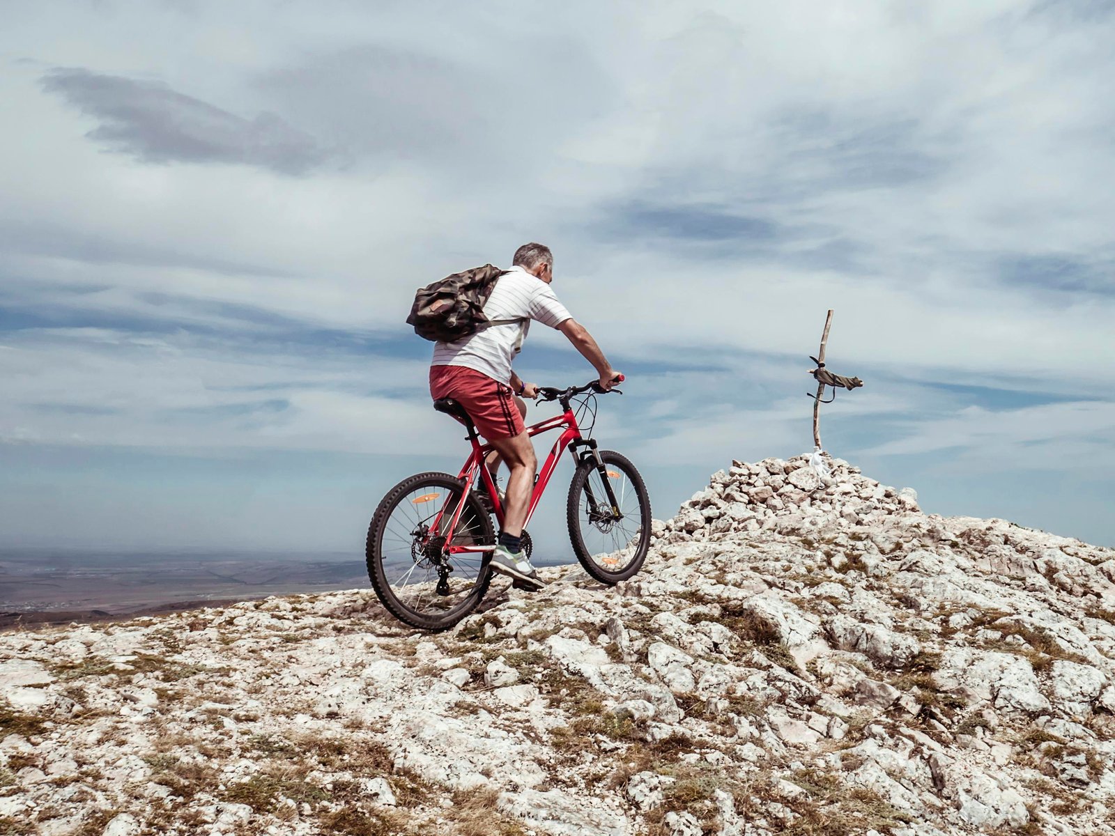 An elderly man biking uphill on a rocky mountain trail under a cloudy blue sky.