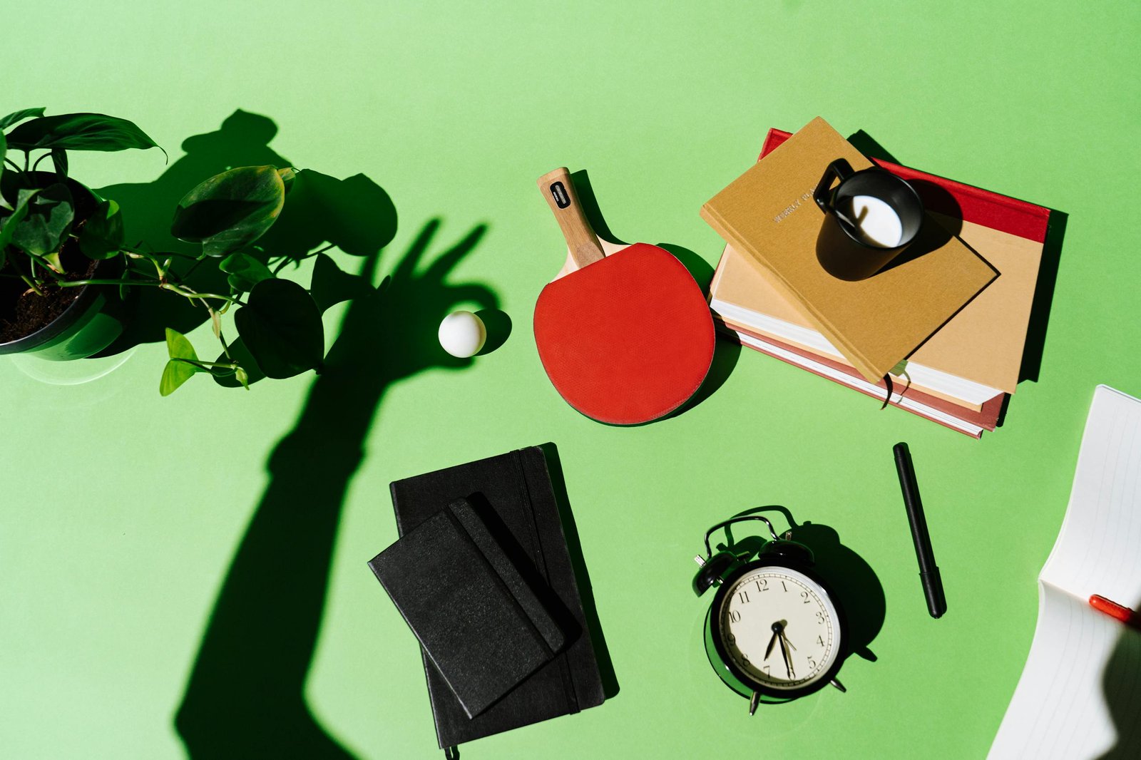 Artistic flat lay of desk items with shadow play, featuring books, a clock, and a plant.