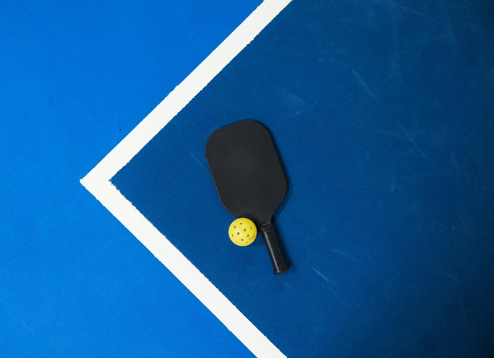 Close-up of a pickleball paddle and ball on a vibrant blue court surface showing geometric lines.