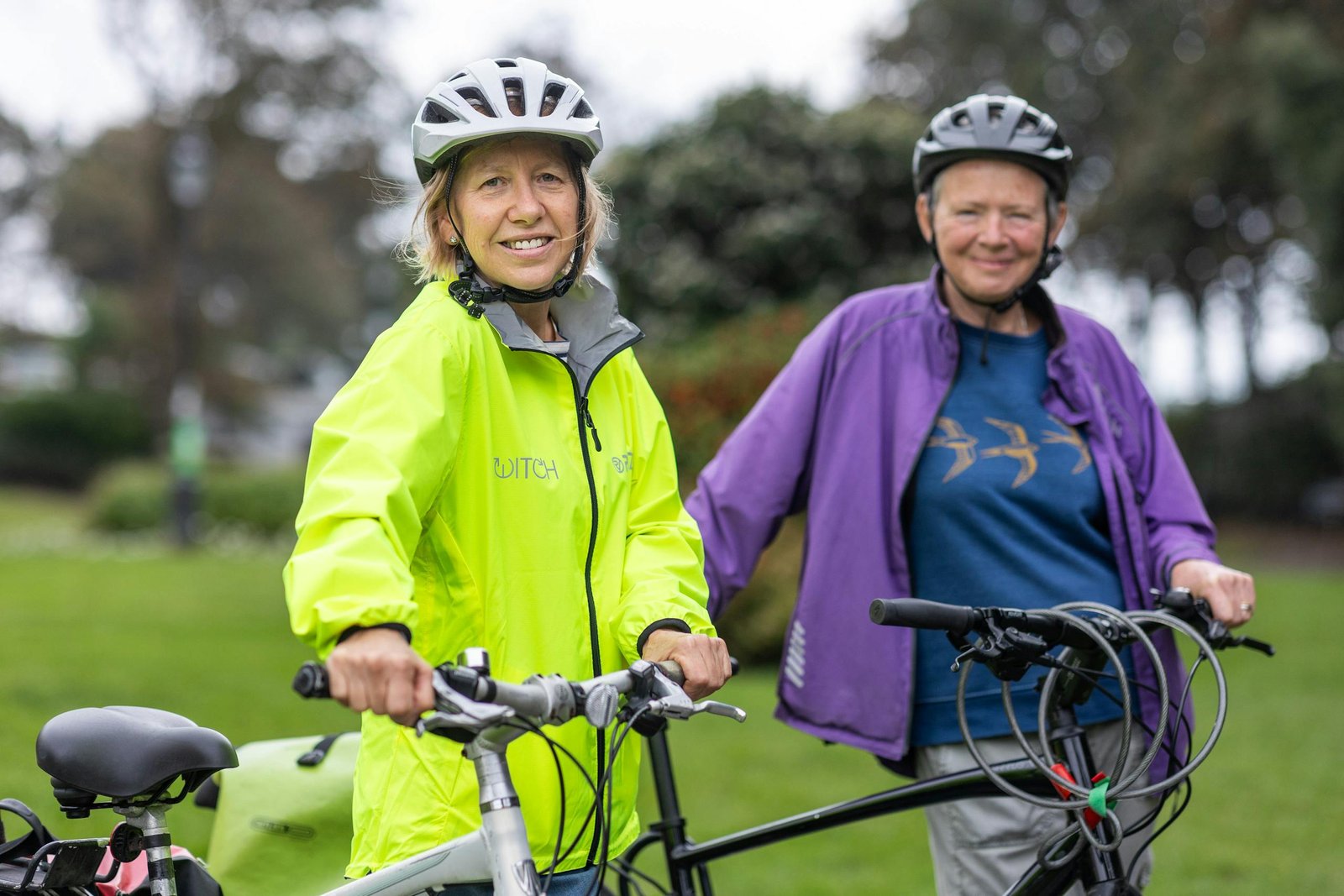 Two women in helmets and bright jackets biking in a park with a blurred natural background.