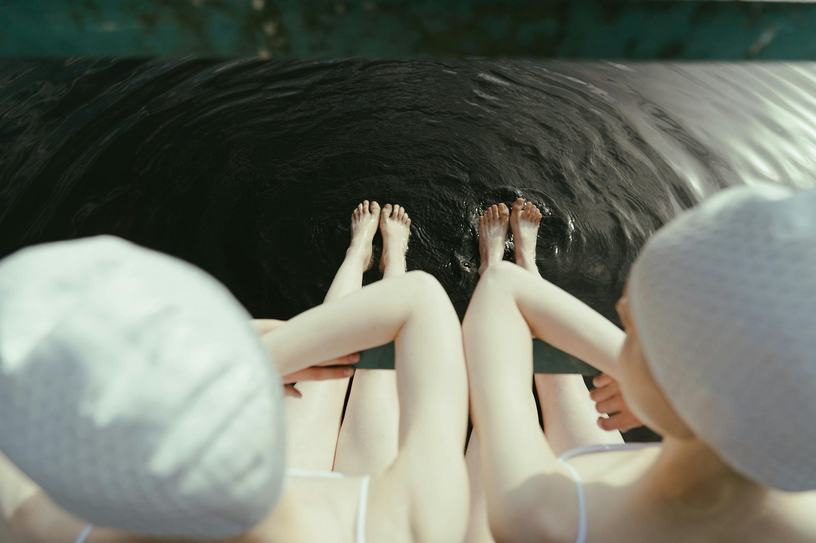 Two women sit by the water's edge, feet dipped in, wearing swim caps and swimwear.