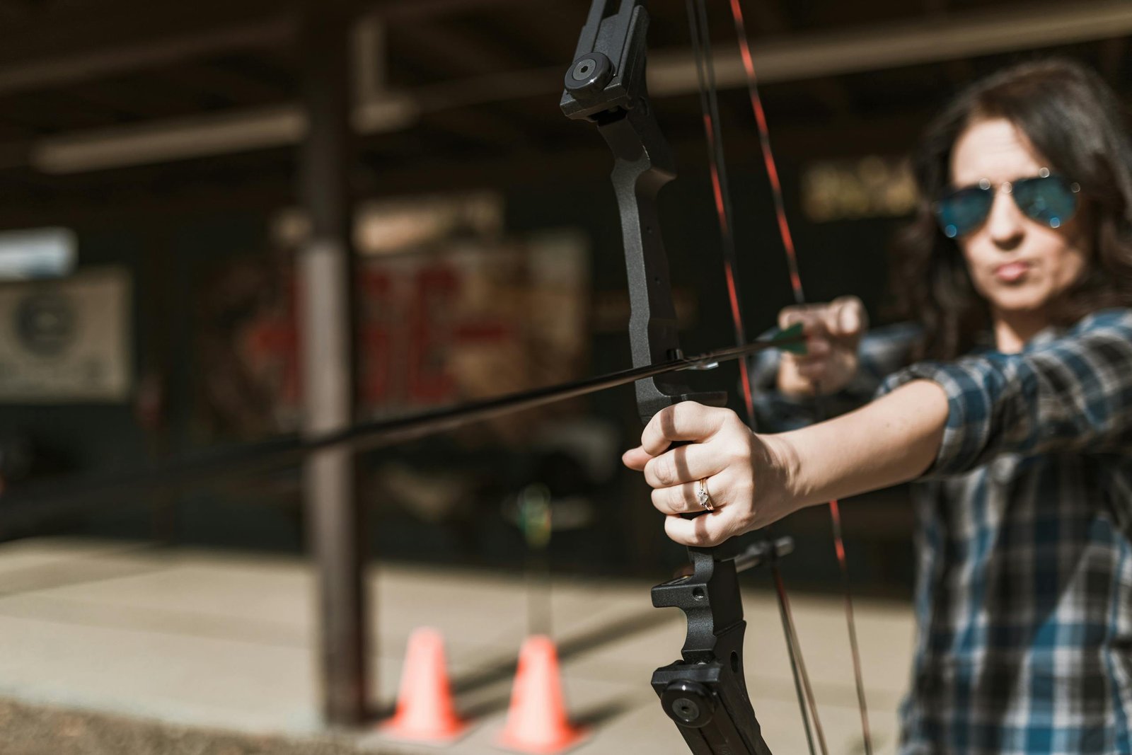 Woman with sunglasses aiming a compound bow outdoors, showcasing concentration and precision.